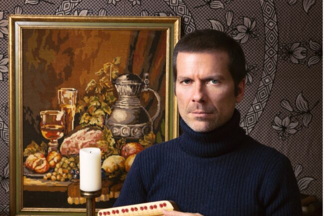 A man in a navy blue turtleneck holds a small wooden keyed instrument while seated in front of a framed still life painting.